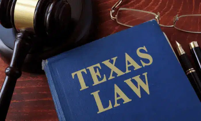 A blue book titled “Texas Law” on a wooden desk, next to a judge’s gavel, eyeglasses, and two pens