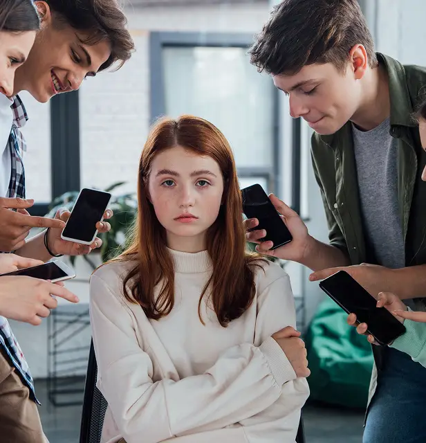 Smiling teenagers pointing with fingers at girl during bullying and holding smartphones with blank