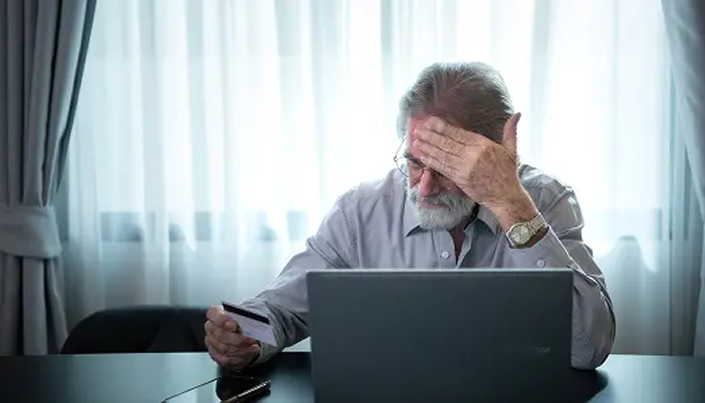 older white man looking at credit card in front of laptop in frustration