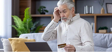 An elderly man sitting at a desk, looking stressed or worried, with one hand resting on his forehead