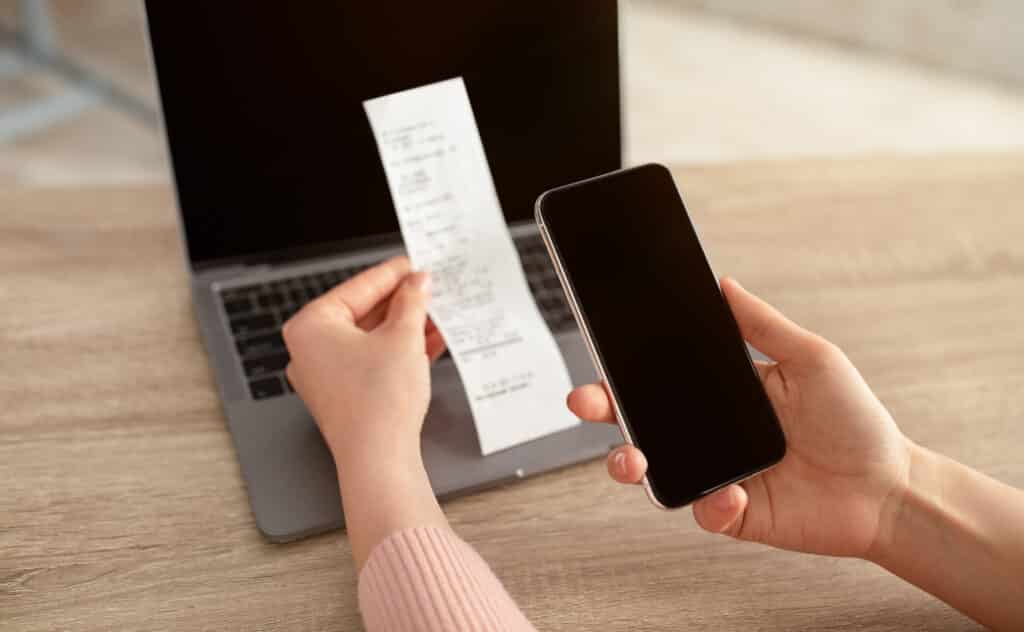 Female hands holding smartphone with empty screen and bill or slip, sitting at workplace with laptop on desk, online payment