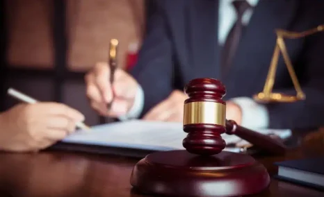 Gavel on a desk with legal professionals reviewing and signing documents in the background, with scales of justice nearby.