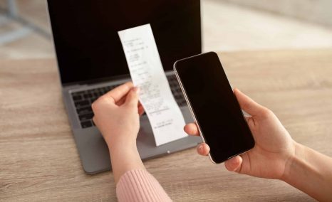 Female hands holding smartphone with empty screen and bill or slip, sitting at workplace with laptop on desk, online payment
